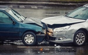 Two cars collided head-on in the middle of a wet road, showing front-end damage and scattered debris.