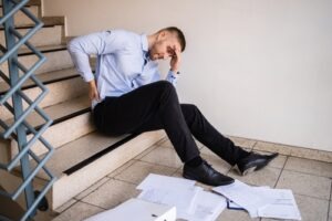Injured man sitting on stairs holding his lower back in pain after a fall, with scattered papers around him.