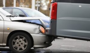 Close-up of a rear-end car collision showing the front of a silver sedan damaged after hitting the back of a gray van on a city street.