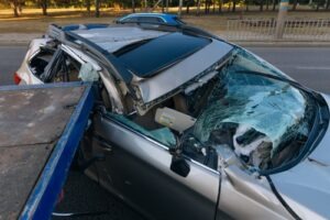 A heavily damaged silver car with a shattered windshield and crushed roof being loaded onto a tow truck after a serious accident.