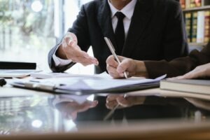 Lawyer meeting with a client at a desk, reviewing and signing legal documents with books and files in the background.