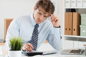 A focused businessman wearing glasses calculates expenses at his desk, reviewing financial documents beside a laptop and small office plant.