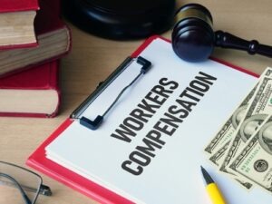 Clipboard labeled “Workers Compensation” on a desk with cash, a gavel, books, and eyeglasses beside it.